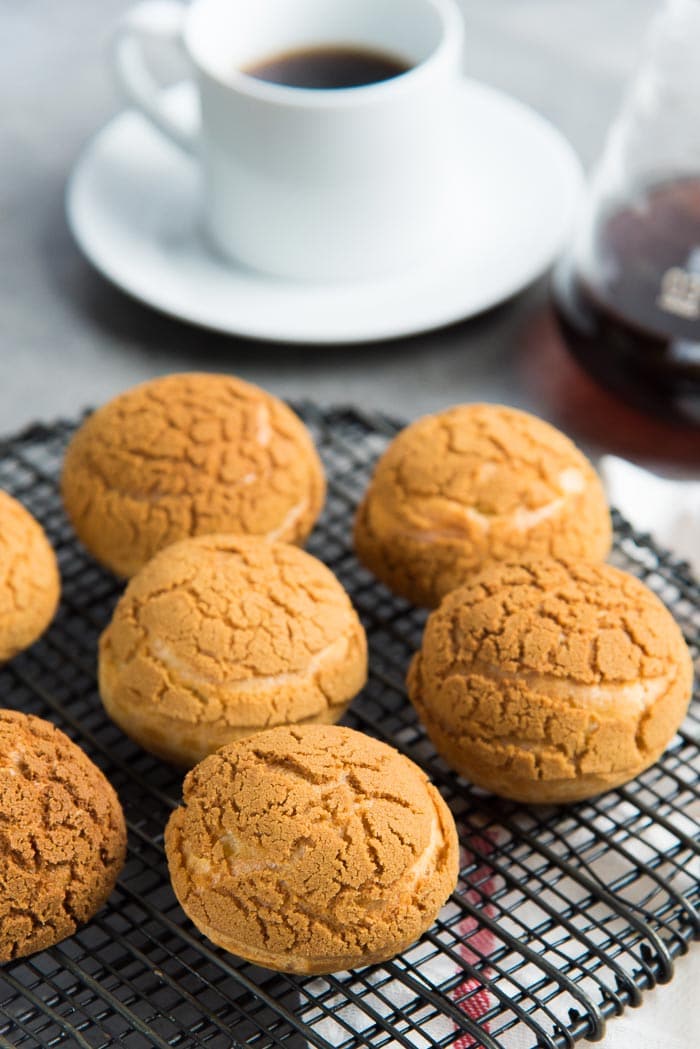 Choux au craquelin with salted caramel cream, placed on top of a black wire rack, with a coffee cup and coffee jug in the background.