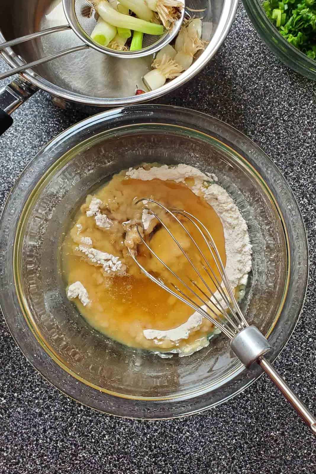 Golden colored oil added to the flour mixture, with a whisk halfway through mixing the contents in the bowl.