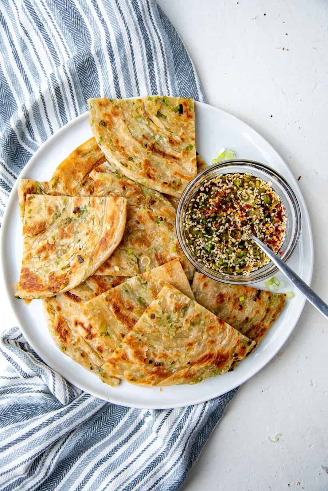 An overhead view of a white plate, with cut up scallion pancakes, and a bowl of soy sauce dipping sauce with sesame seeds floating.