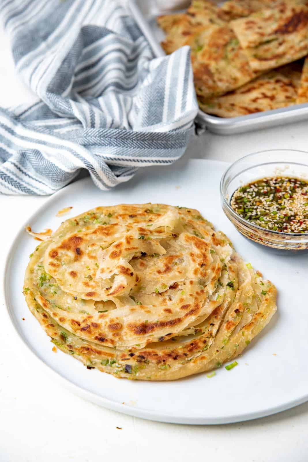 Two whole scallion pancakes placed on a while plate with a blue kitchen towel and the dipping sauce in the background.