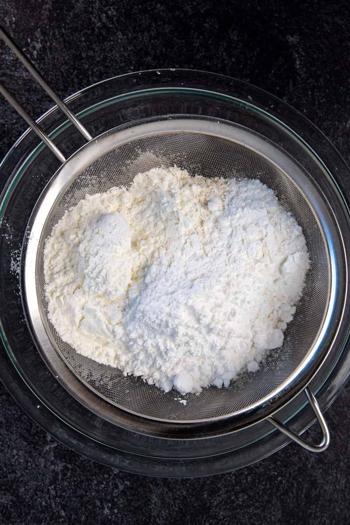 A sieve placed ontop of a large glass bowl, with all the dry ignredients placed in the sieve.
