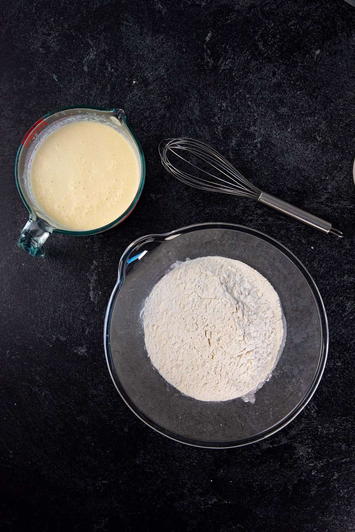 An overhead view of the wet ingredients that are mixed in a separate jug, a large bowl with the mixed dry ingredients separately and a whisk next to them.