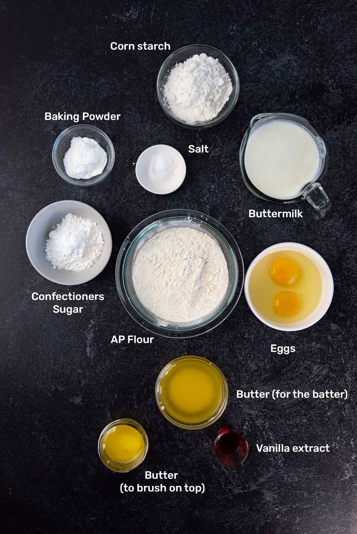Overhead view of labeled ingredients needed to make sheet pan pancakes from scratch. From top (clockwise), cornstarch, buttermilk, eggs, vanilla extract, butter for the batter, butter to brush on top, confectioners sugar, baking powder, salt and AP flour (in the center).