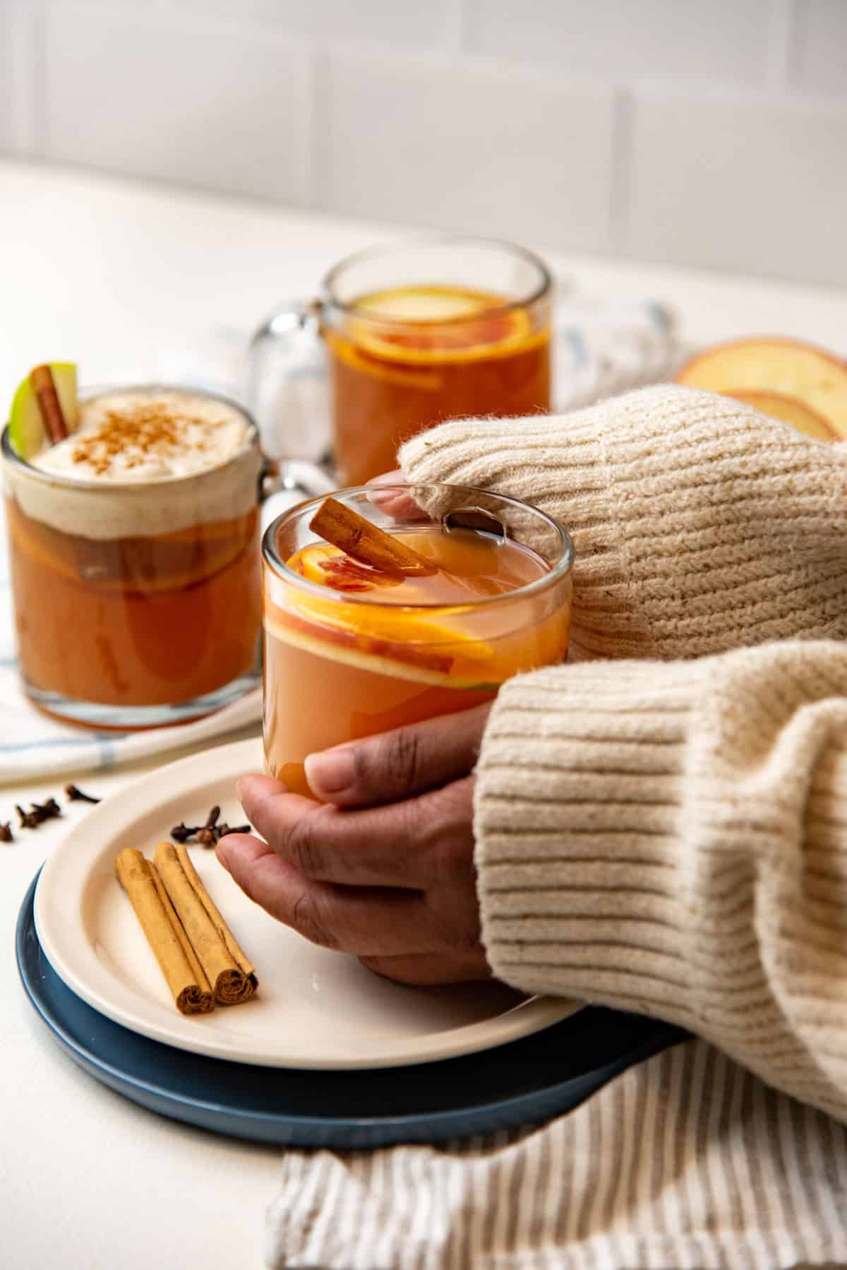 Two hands holding a glass mug of spiced warm apple cider, with two mugs in the background.