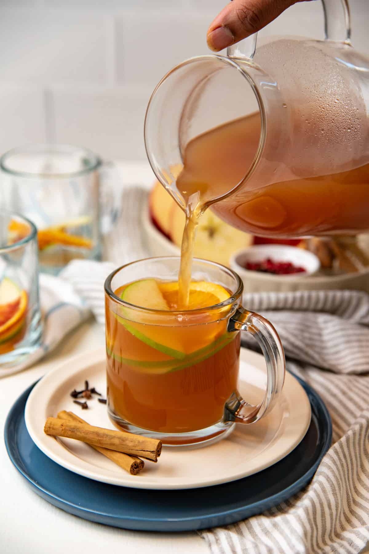 Pouring warm spiced apple cider into a glass mug with slices of apples and oranges in the mug as garnish.