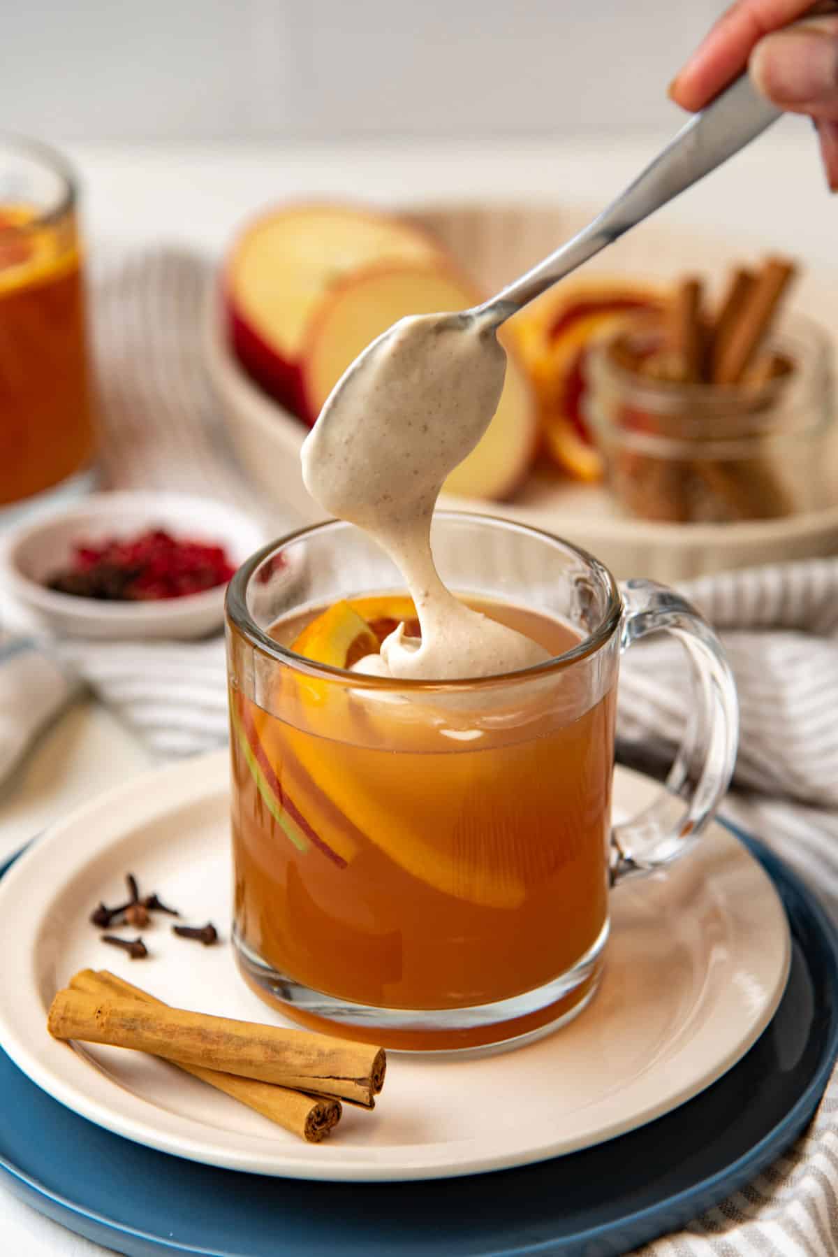 A spoonful of cinnamon whipped cream being placed ontop of warm apple cider.