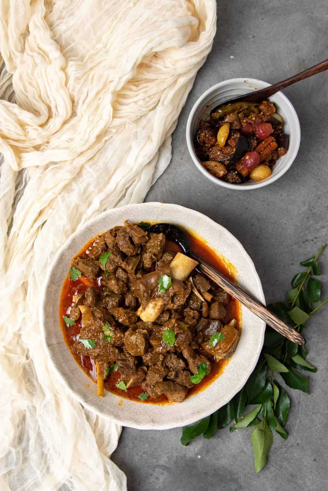 Overhead view of mutton curry with bones, and an eggplant pickle in a separate bowl.