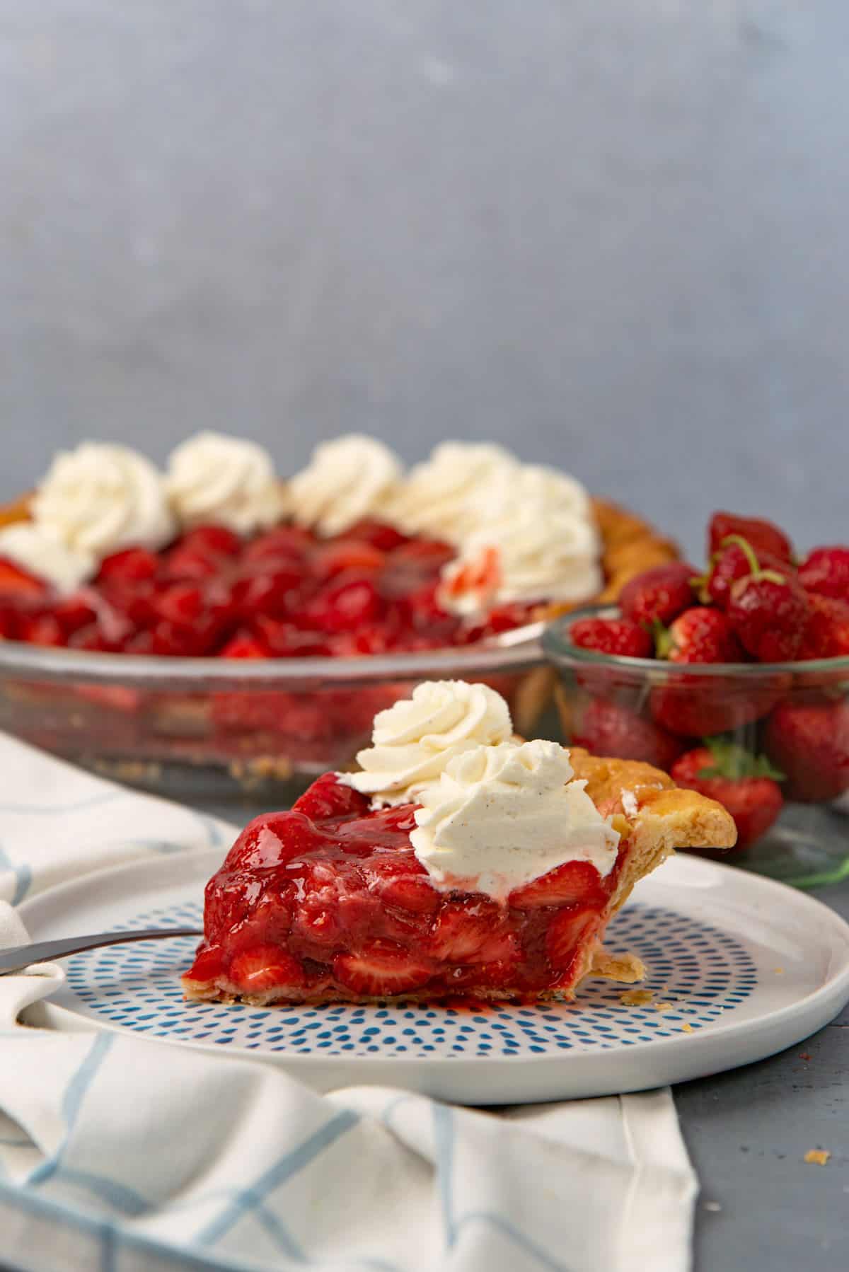 Fresh strawberry pie slice with whipped cream swirls on top and bowl of strawberries in background.