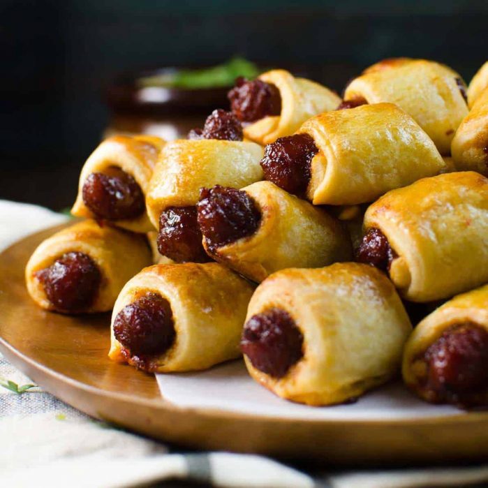 Wooden platter with a big stack of pigs in a blanket, showing the flaky pastry and juicy sausages.