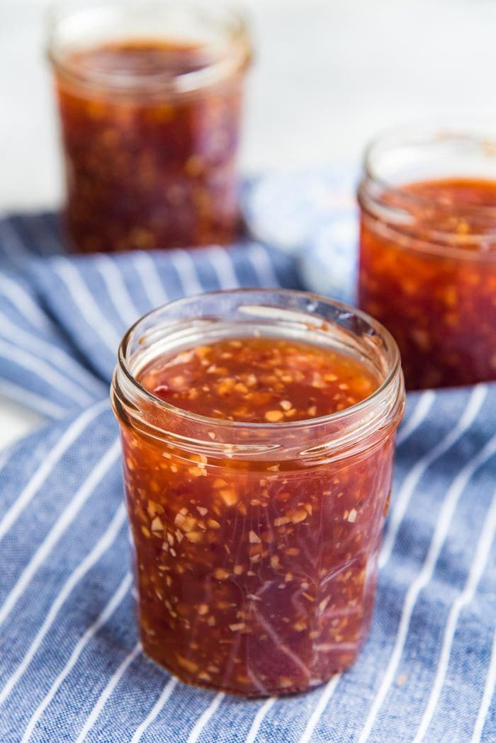 The BEST Homemade Sweet Chili Sauce in a jar, ready to eat or be canned, on a cloth napkin. With two more jars in the background.