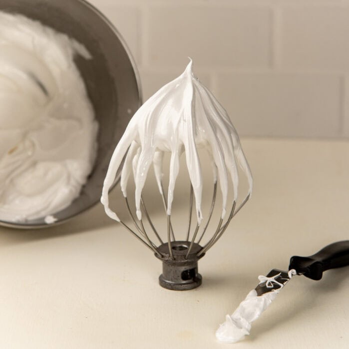 Swiss meringue with stiff peaks on a whisk, with a bowl of meringue in the background.