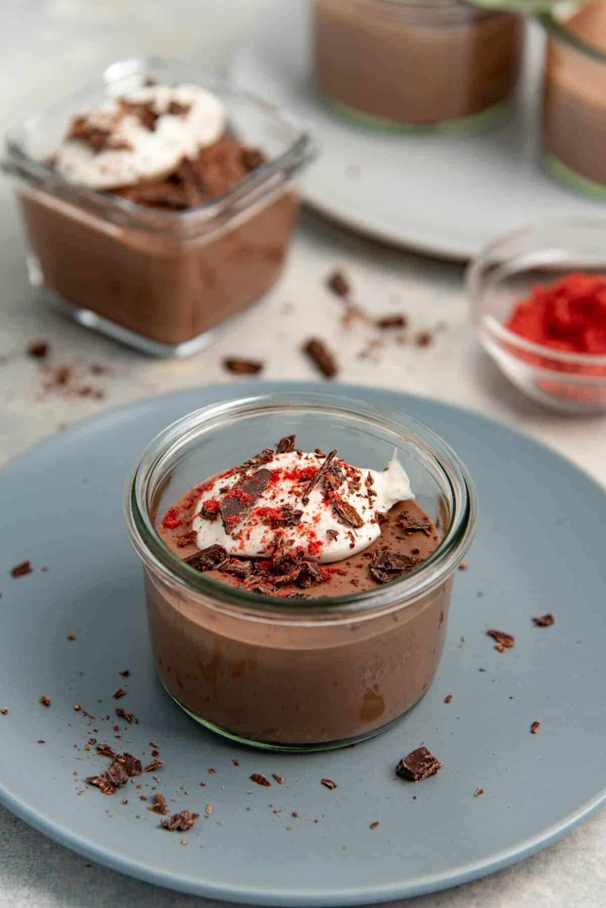 Tofu chocolate mousse in a round glass container, topped with whipped cream, chocolate shavings and raspberry powder.