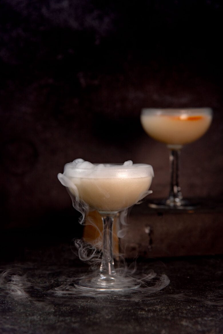 A martini glass with a peach colored drink, silvery white shimmery swirls, and smoke from dry ice.